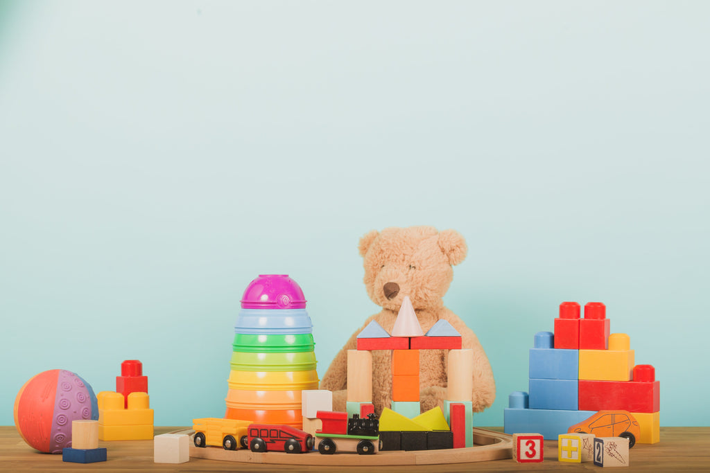 I notice there's a discrepancy - the image shows a teddy bear with toys, not a "Baby Alien Plushie." Based on what's actually visible:

Tan teddy bear surrounded by colorful toys including blocks, stacking rings, wooden train, and ball on light blue background.

However, if you need alt text specifically for a "Baby Alien Plushie" product, please provide the correct image.